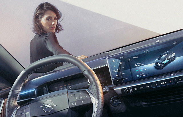 Eine Frau steht draußen neben einem Auto und schaut über ihre Schulter in die Kamera. Im Vordergrund ist das Innere eines Autos sichtbar, einschließlich des Lenkrads mit Opel-Logo und eines modernen digitalen Armaturenbretts, das verschiedene Anzeigen zeigt.