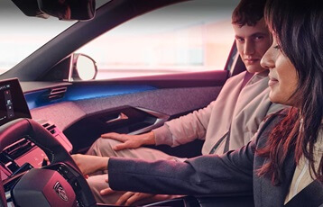 Zwei Personen sitzen in einem Auto, die Frau auf dem Fahrersitz und der Mann auf dem Beifahrersitz. Die Frau hält ihre Hand auf dem Armaturenbrett, während der Mann in ihre Richtung schaut. Das Innere des Autos ist modern gestaltet mit einem digitalen Display und einer blauen Beleuchtung.