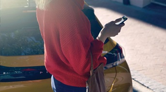 Eine Person steht neben einem gelben Auto und hält ein Smartphone in der Hand. Die Person trägt einen roten Pullover und eine Tasche über der Schulter. Im Hintergrund ist eine gepflasterte Straße zu sehen.